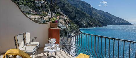 A balcony with a table set for two, a chair, and a yellow cushion. The balcony overlooks a coastal area with buildings on a hillside and the sea.