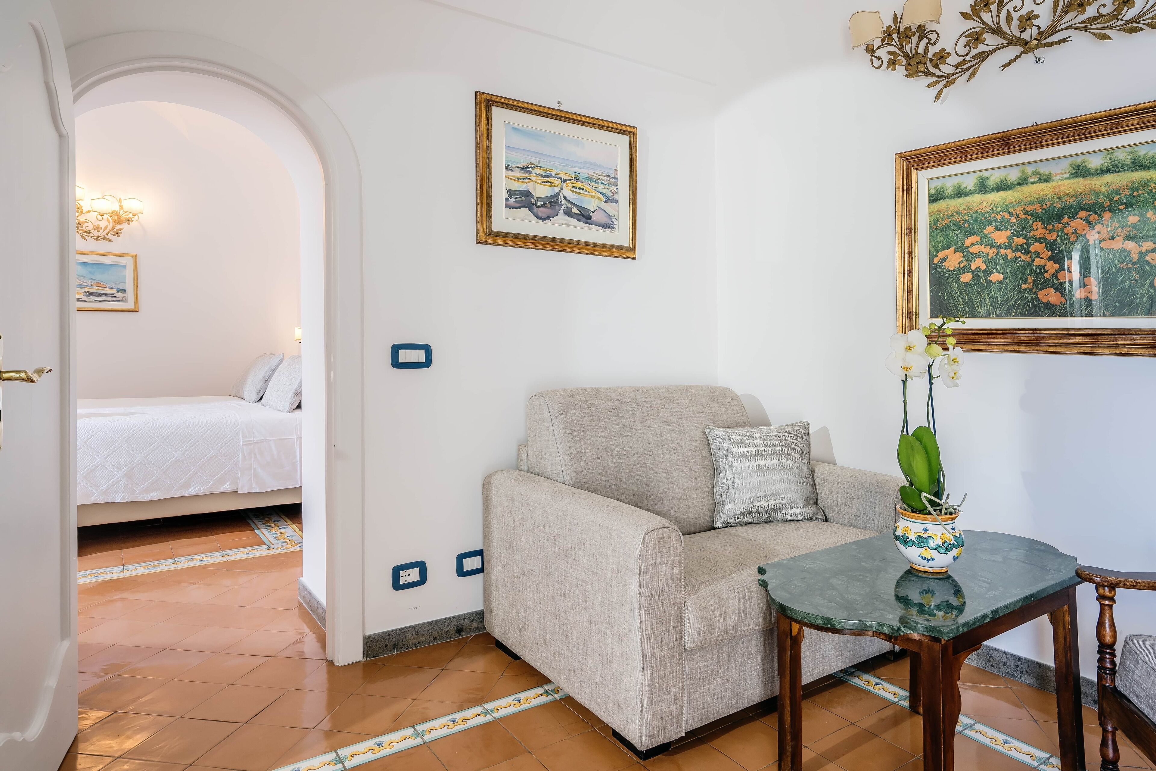A room with a sofa, a small table with a vase, and a bed in the background.