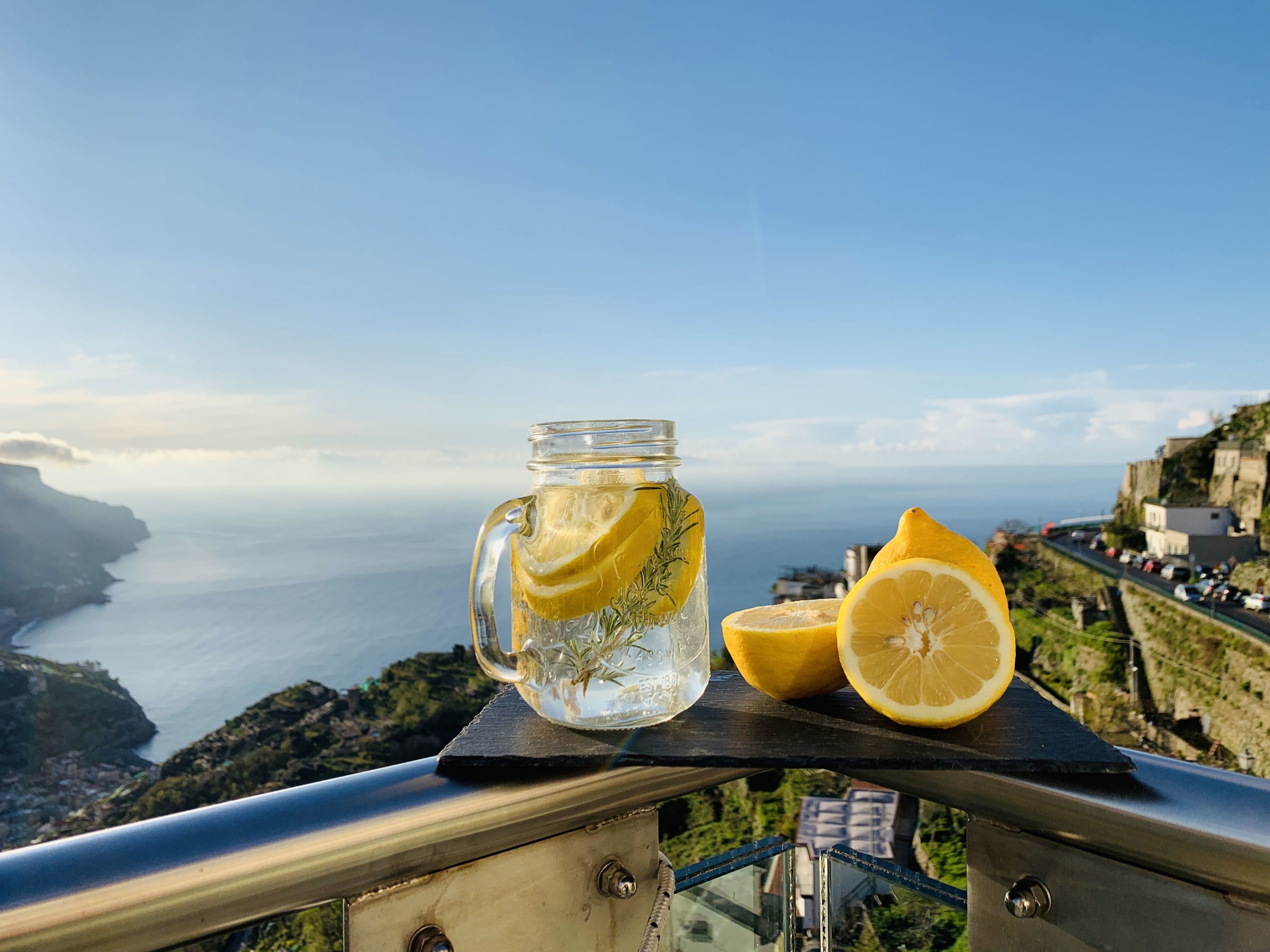 A glass jar with lemon slices and a lemon half on a balcony overlooking a coastal view.
