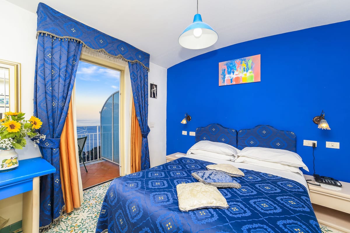 A bedroom with a blue wall, a bed with patterned bedding, a nightstand, a lamp, and a view of the outdoors.