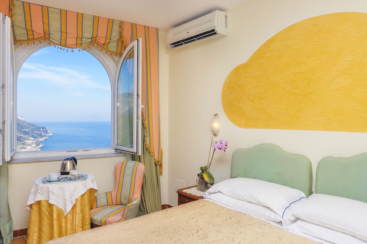 A hotel room with a bed, a chair, a small table with a tray, a window with curtains, and a view of the sea.