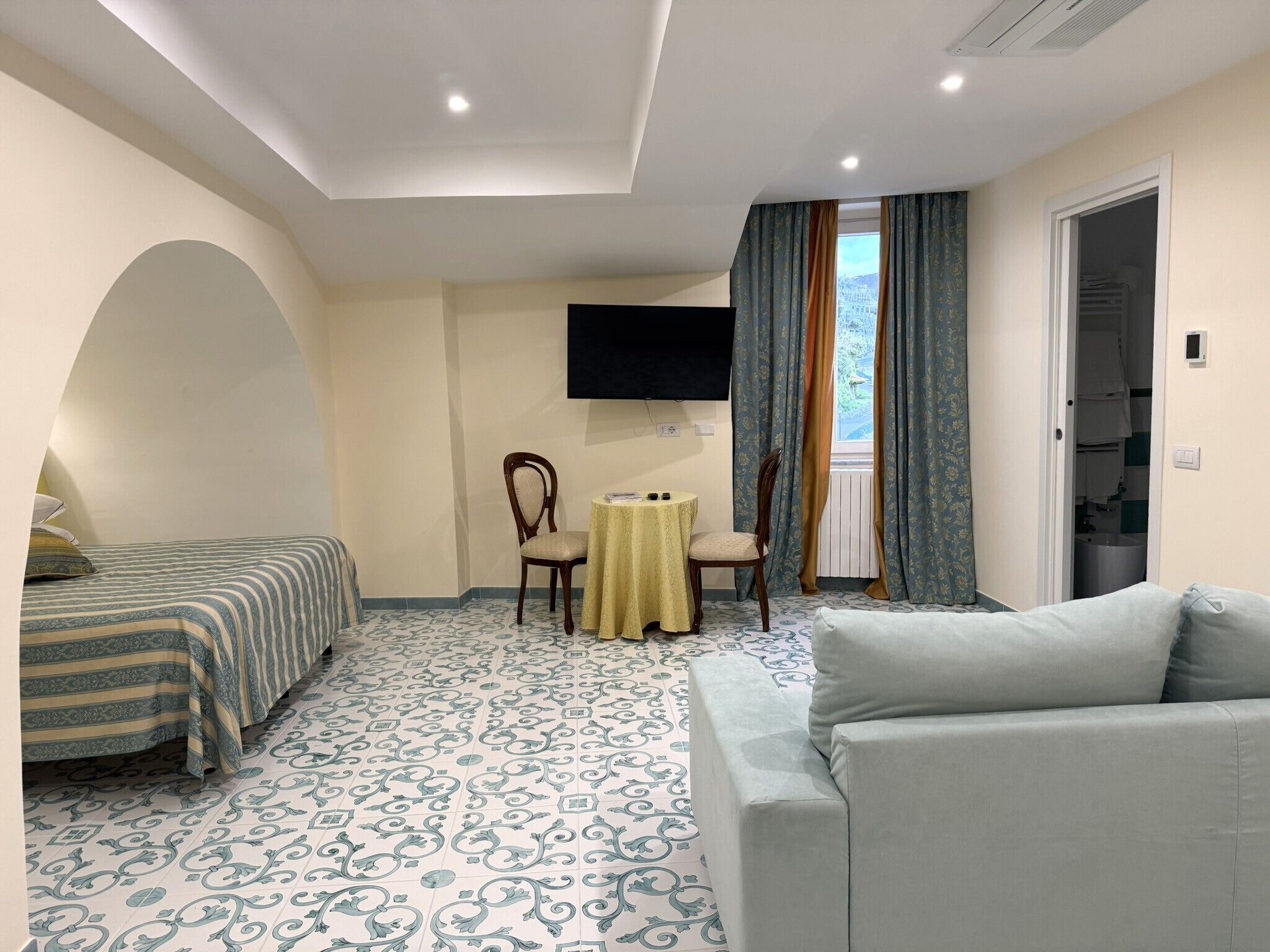 Habitación de hotel con cama, sofá, mesita, sillón, televisor y ventana con cortinas.