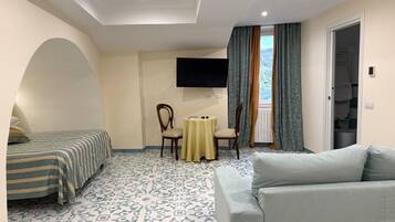 Habitación de hotel con cama, sofá, mesita, sillón, televisor y ventana con cortinas.