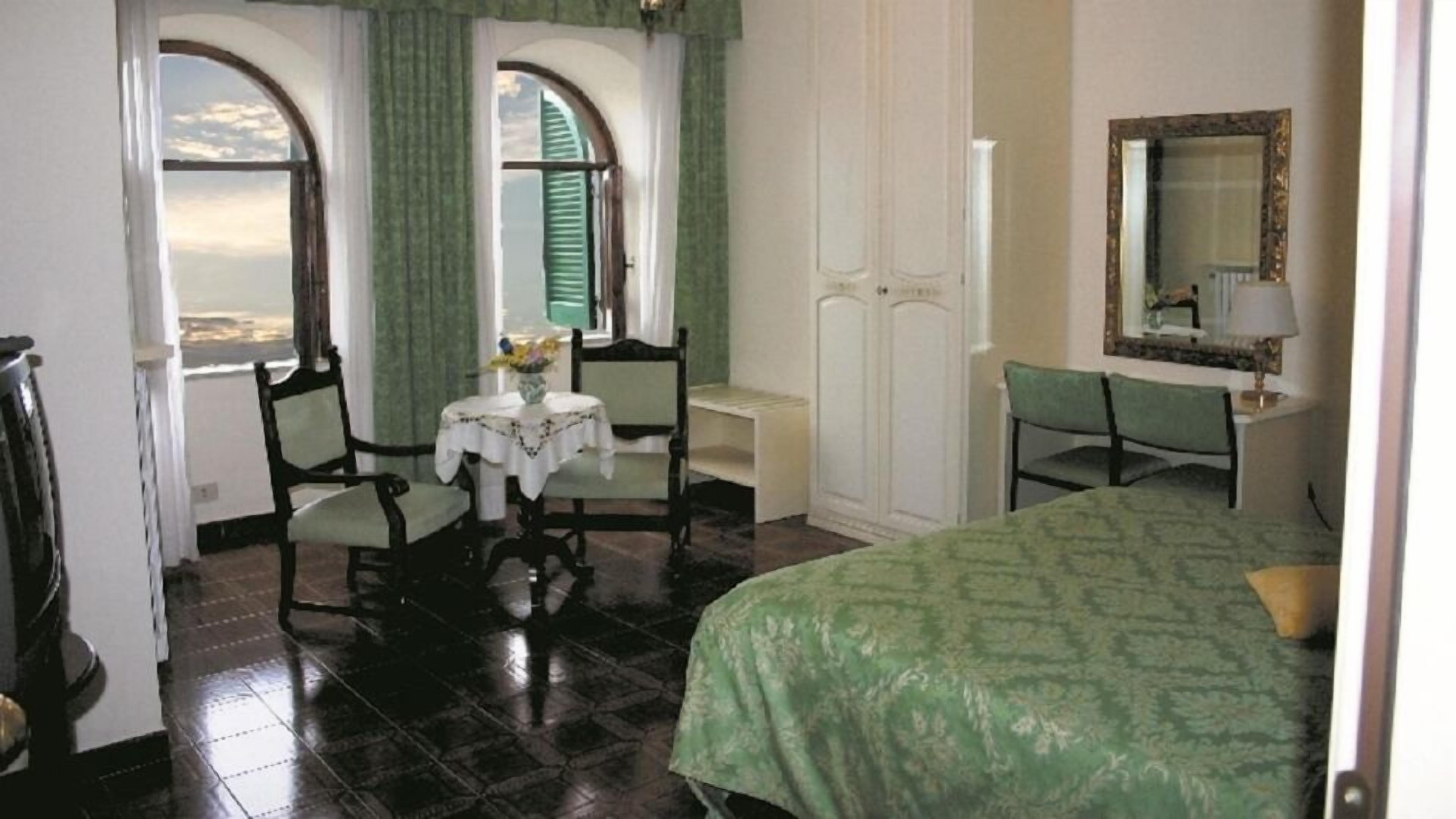 Un dormitorio con cama, escritorio, silla, mesa con un florero y dos ventanas con contraventanas.