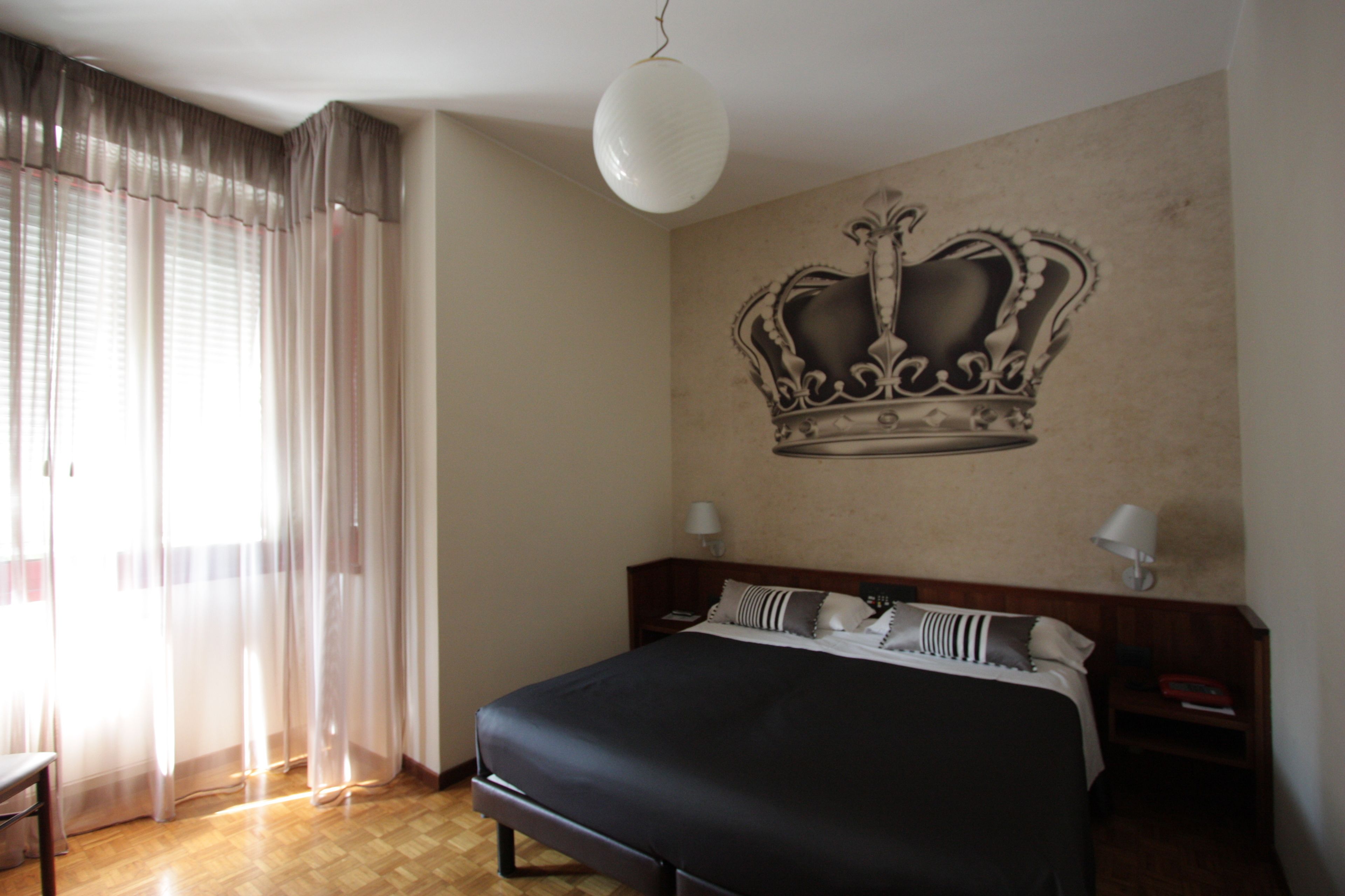 A bedroom with a large bed, a crown mural on the wall, a wooden floor, and a window with sheer curtains.