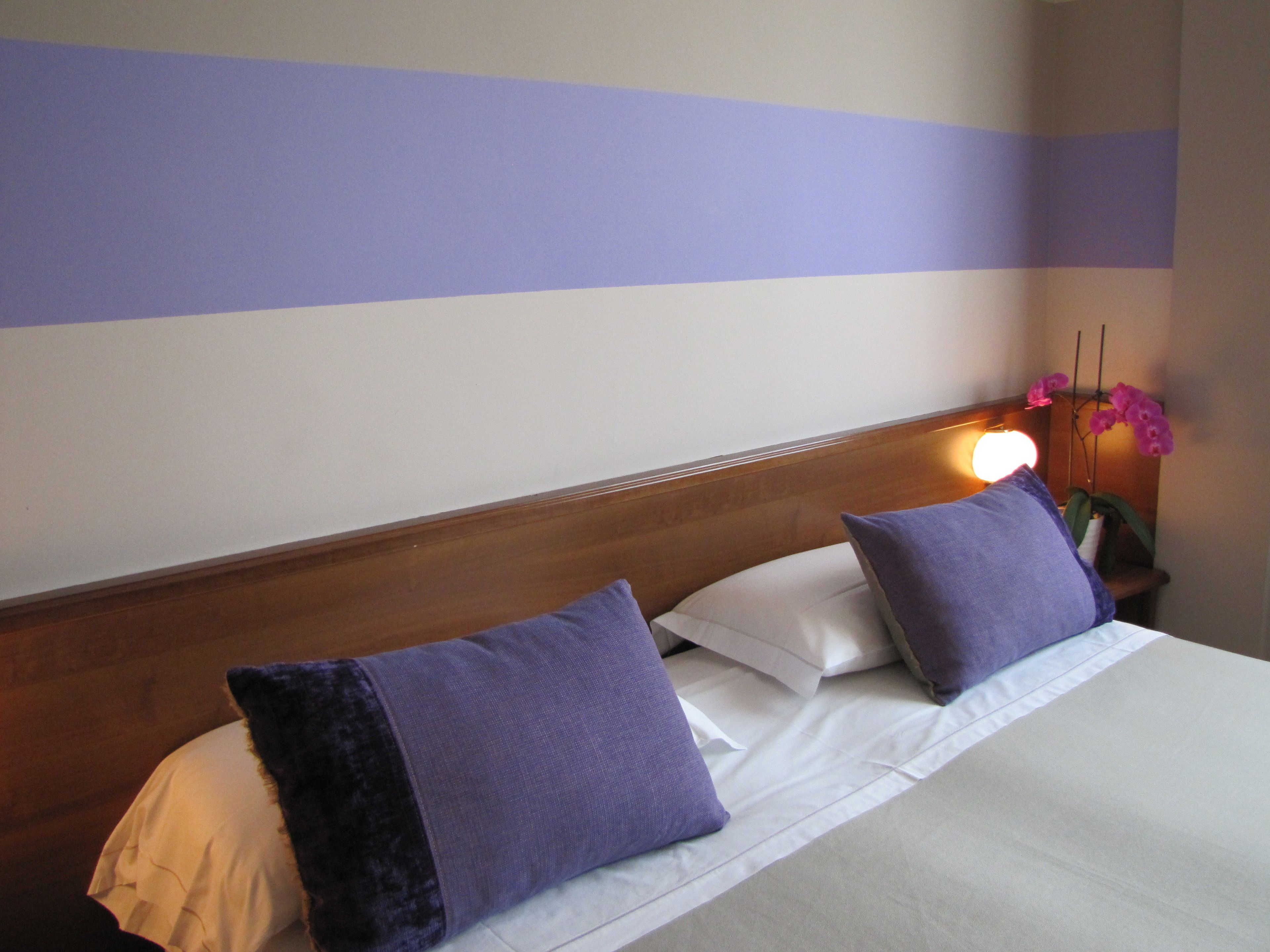 Ein ordentlich bezogenes Bett mit weißen Leinen und zwei violetten Kissen, ein Holz-Kopfstück und eine Wand mit horizontalen Streifen.