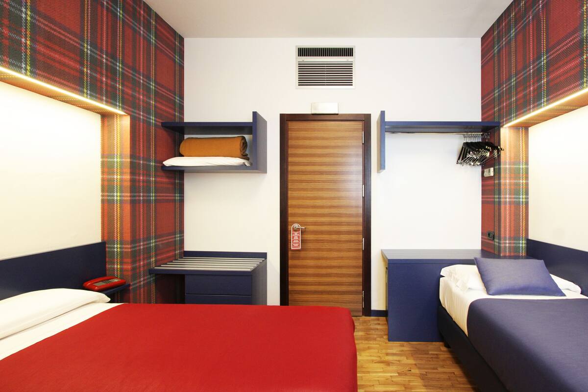A room with a large bed, two smaller beds, a wardrobe, a shelf with towels, and a coat rack.