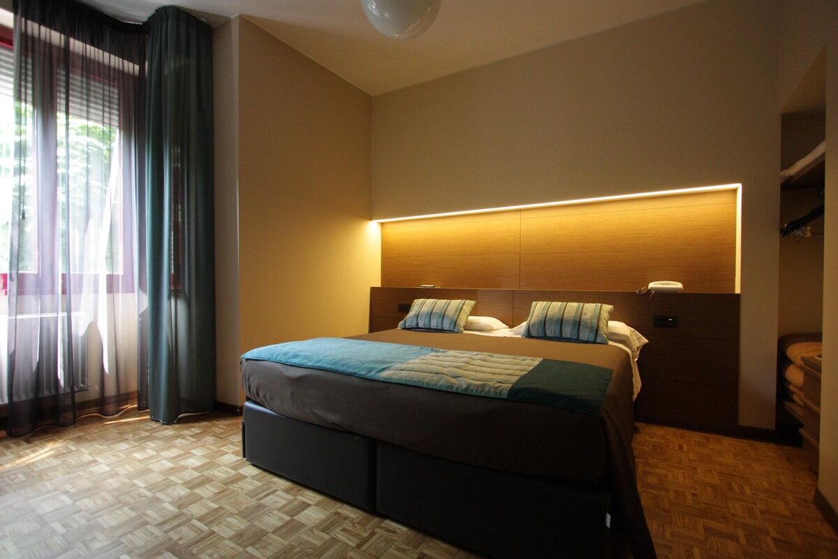 Une chambre d’hôtel moderne dotée d’un grand lit, d’une table de chevet, d’une tête de lit intégrée avec éclairage intégré, d’un plancher en bois et offrant une vue sur l’extérieur à travers des rideaux transparents.