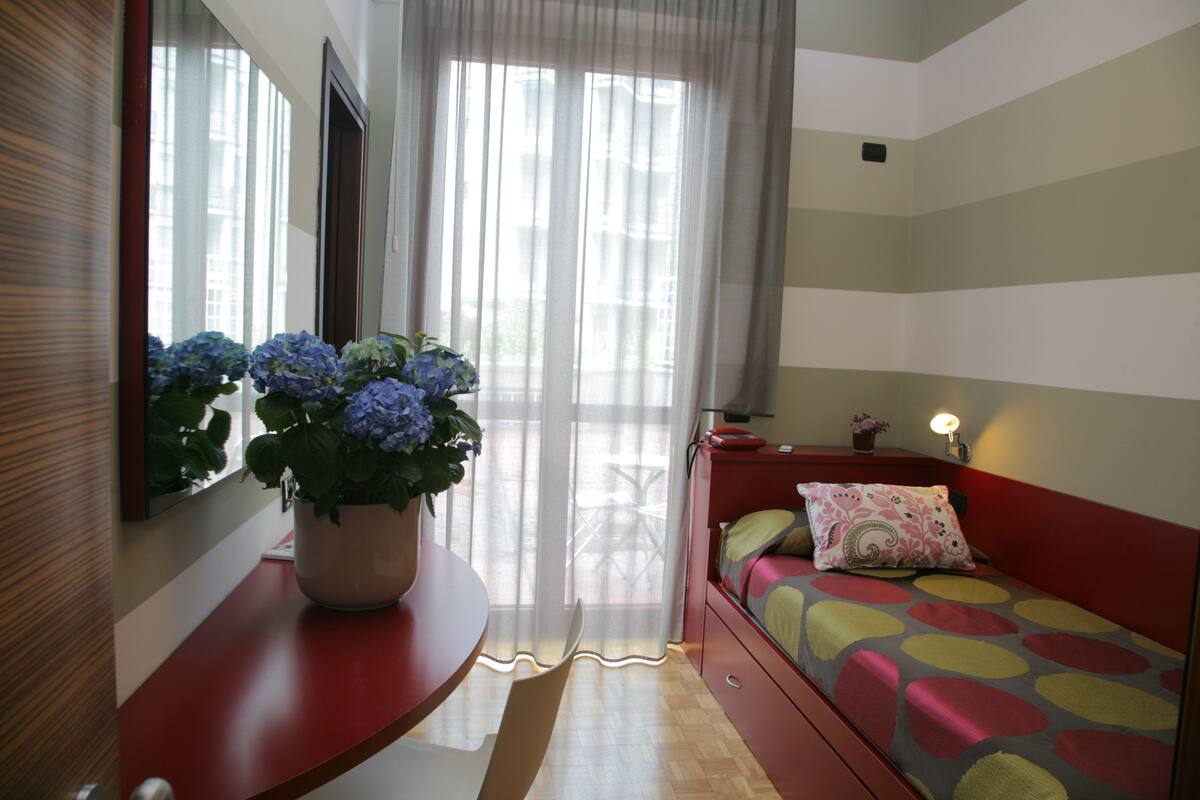 A bedroom with a bed, a bedside table, a mirror, and a window with curtains.