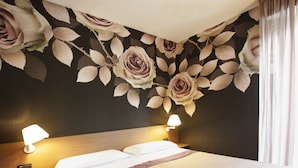 Hotelkamer met een bed, twee bedlampjes, een nachtkastje met een rode wekker en een bloemen behang.