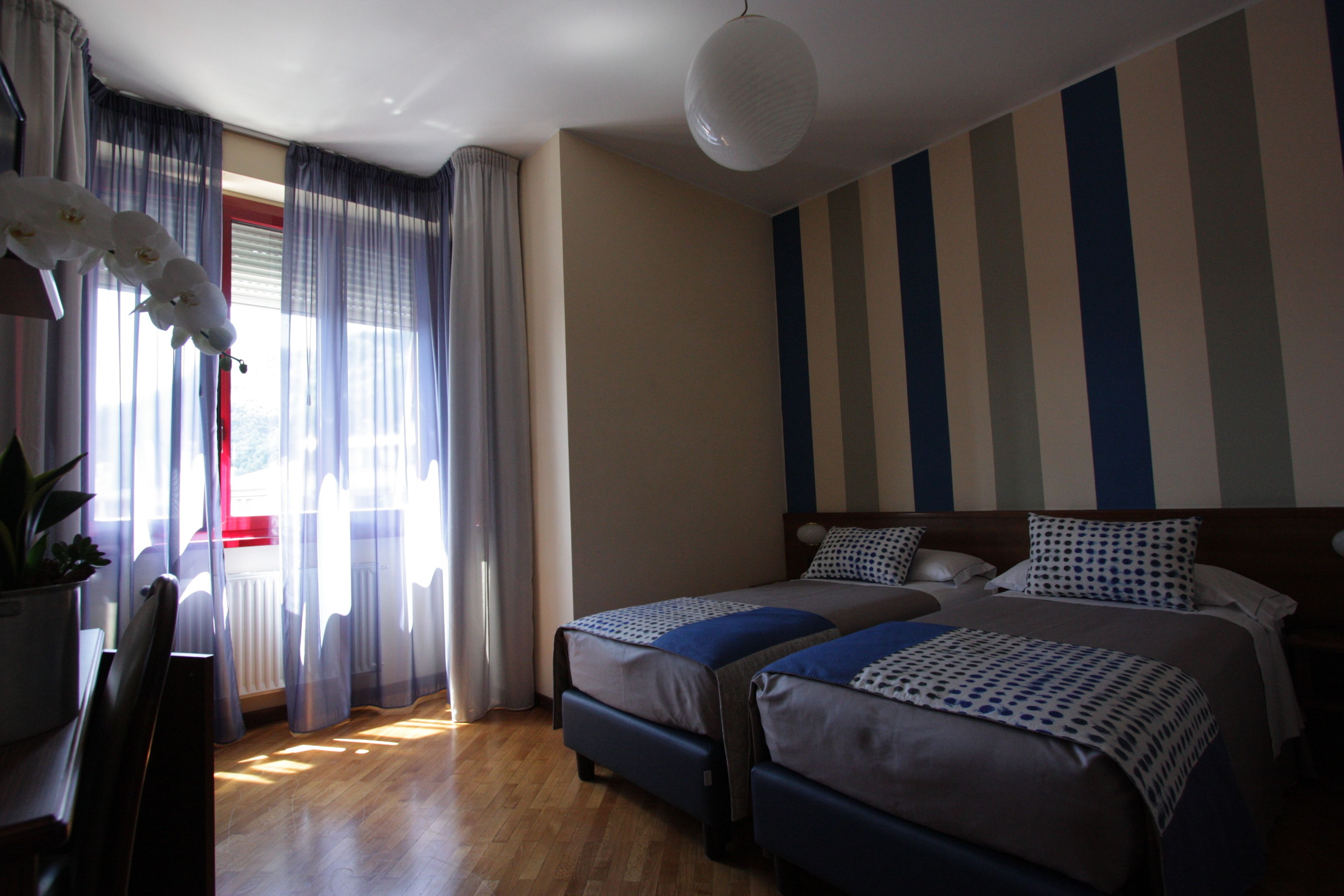 Habitación de hotel con dos camas, una planta en maceta y vistas al exterior a través de cortinas translúcidas.