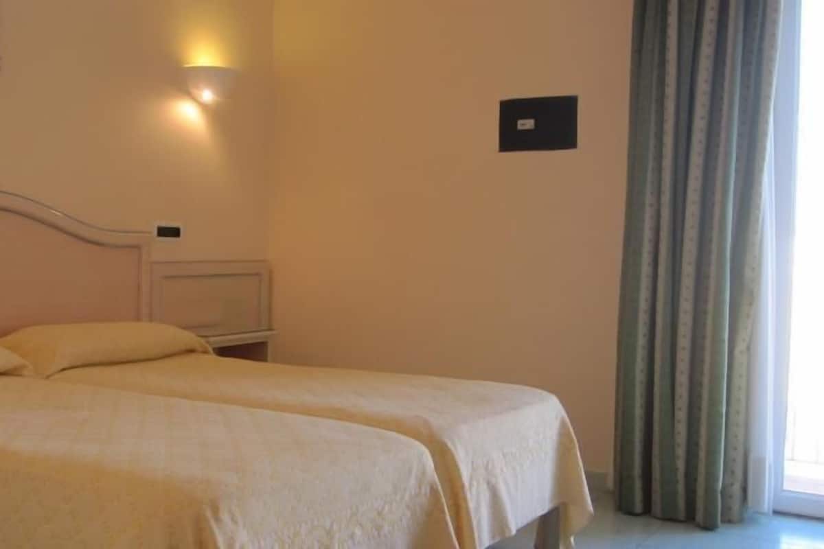 A double bed with two pillows in a room with a nightstand, a wall-mounted light, and a window with curtains.
