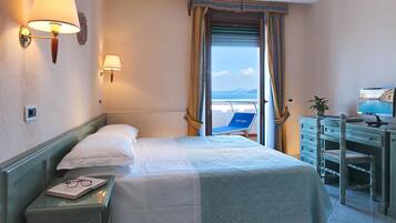A hotel room with a bed, a desk, a chair, a TV, and a view of the sea through the window.
