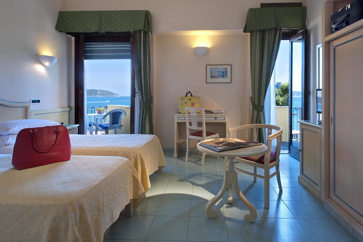 A hotel room with two beds, a desk, a chair, and a view of the sea.
