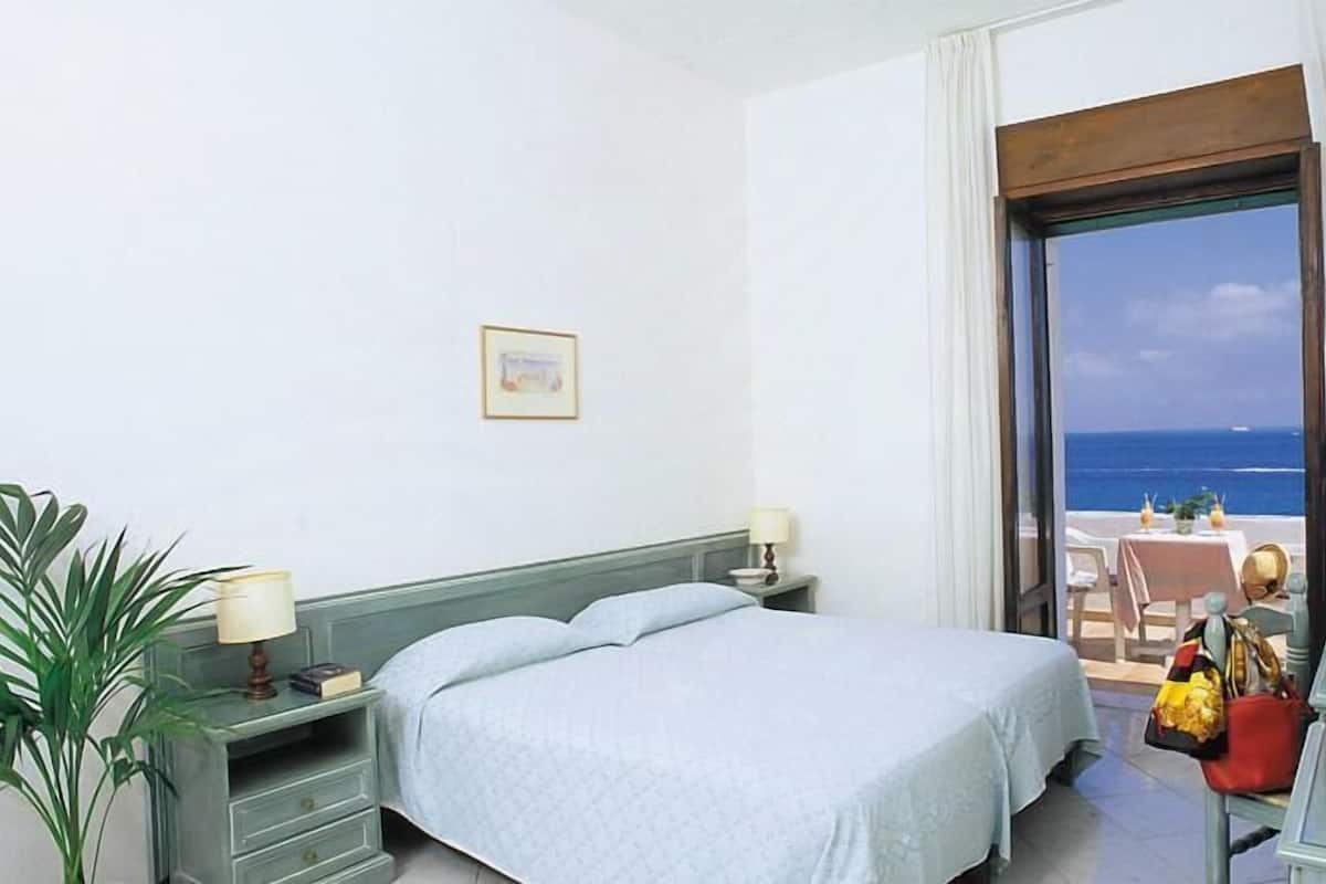 A hotel room with a bed, bedside tables, a desk with a chair, a television, and a view of the sea.