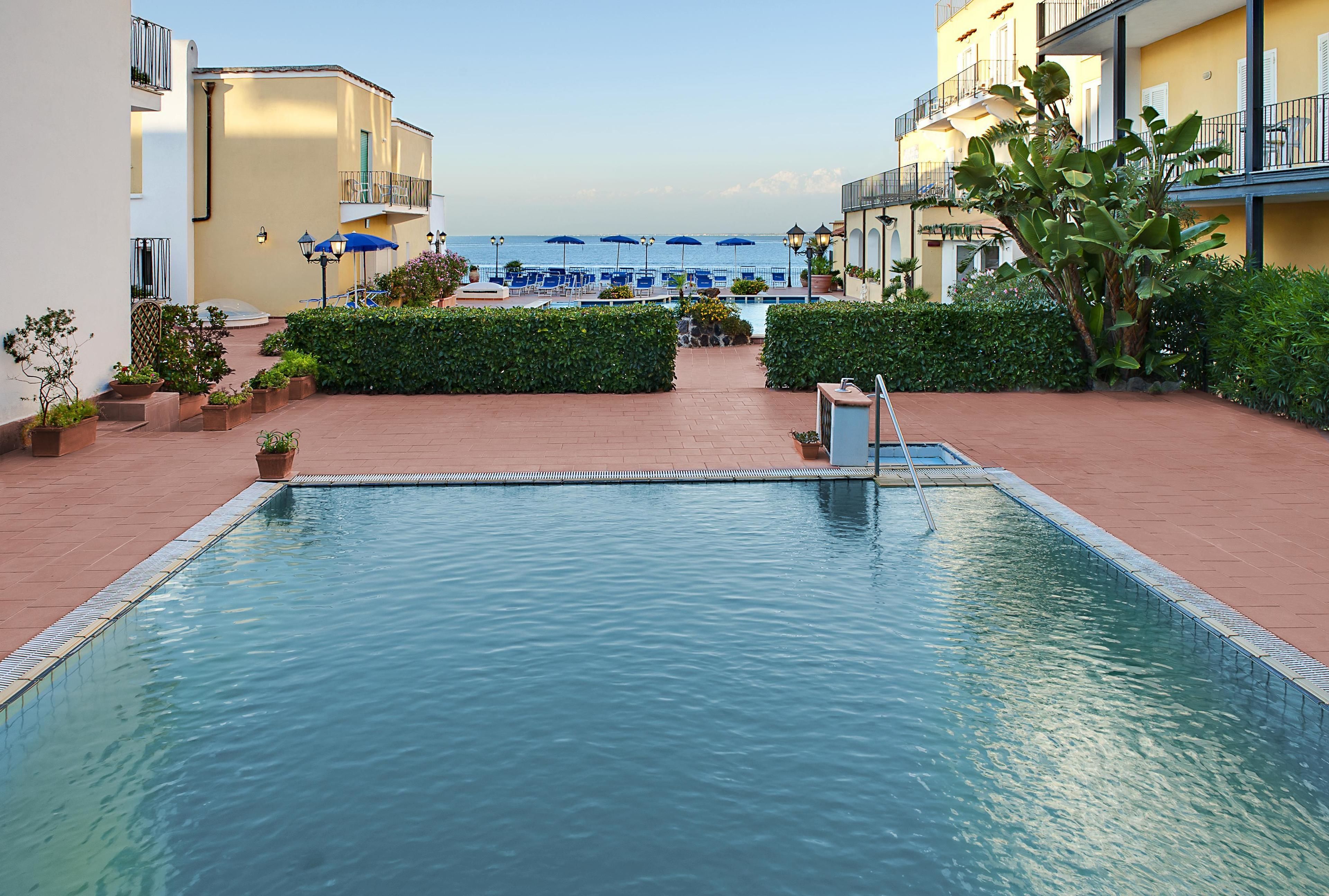 A pool area with a clear view of the sea, surrounded by potted plants and umbrellas.