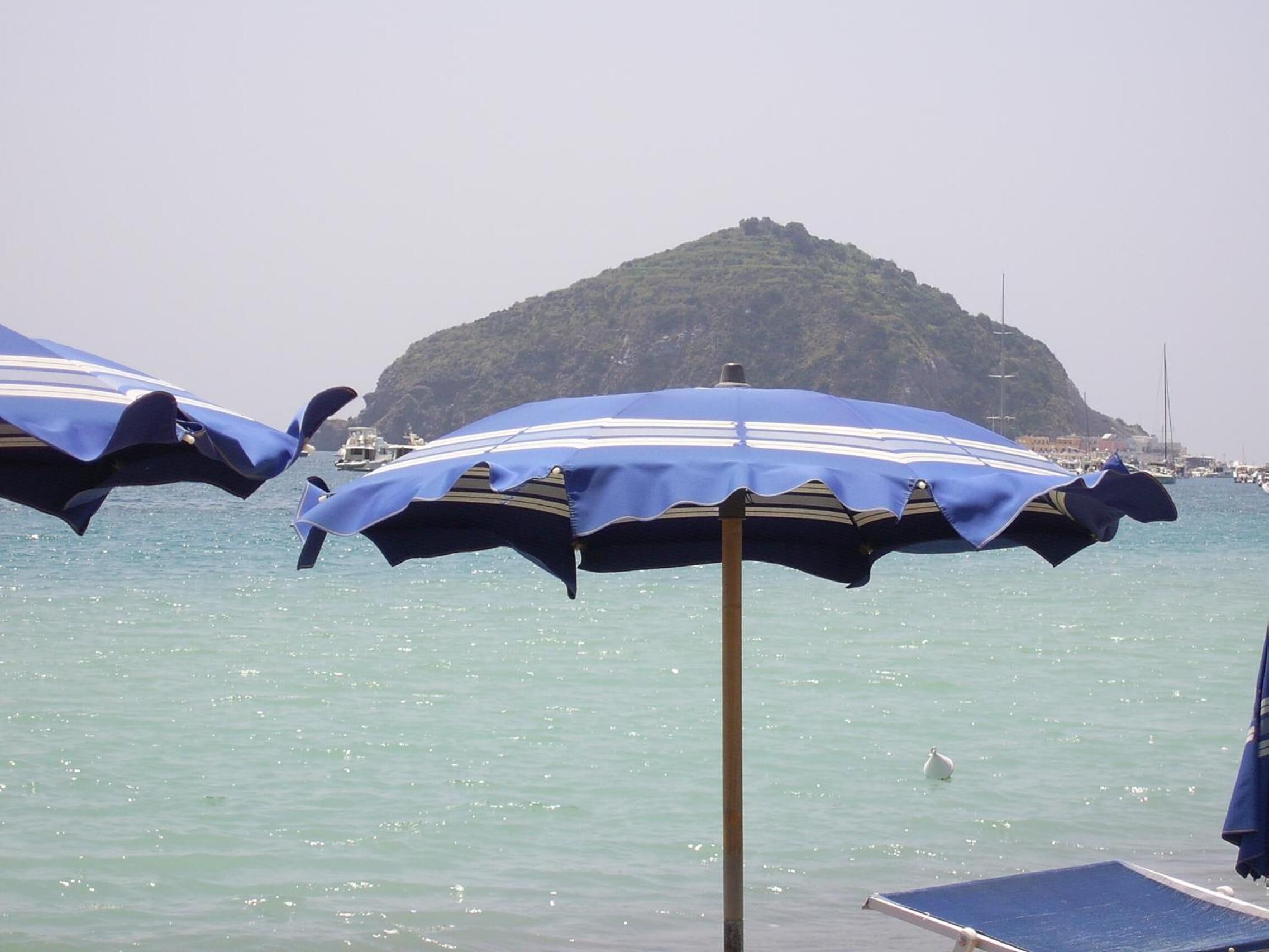 A seaside view with a small island in the background and two blue umbrellas in the foreground.