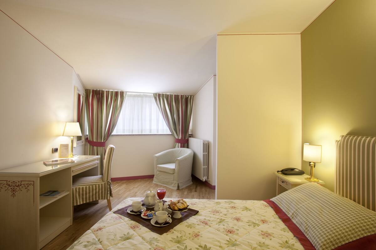 Ein Hotelzimmer mit Bett, Schreibtisch, Stuhl, Heizkörper und einem Fenster mit Vorhängen.