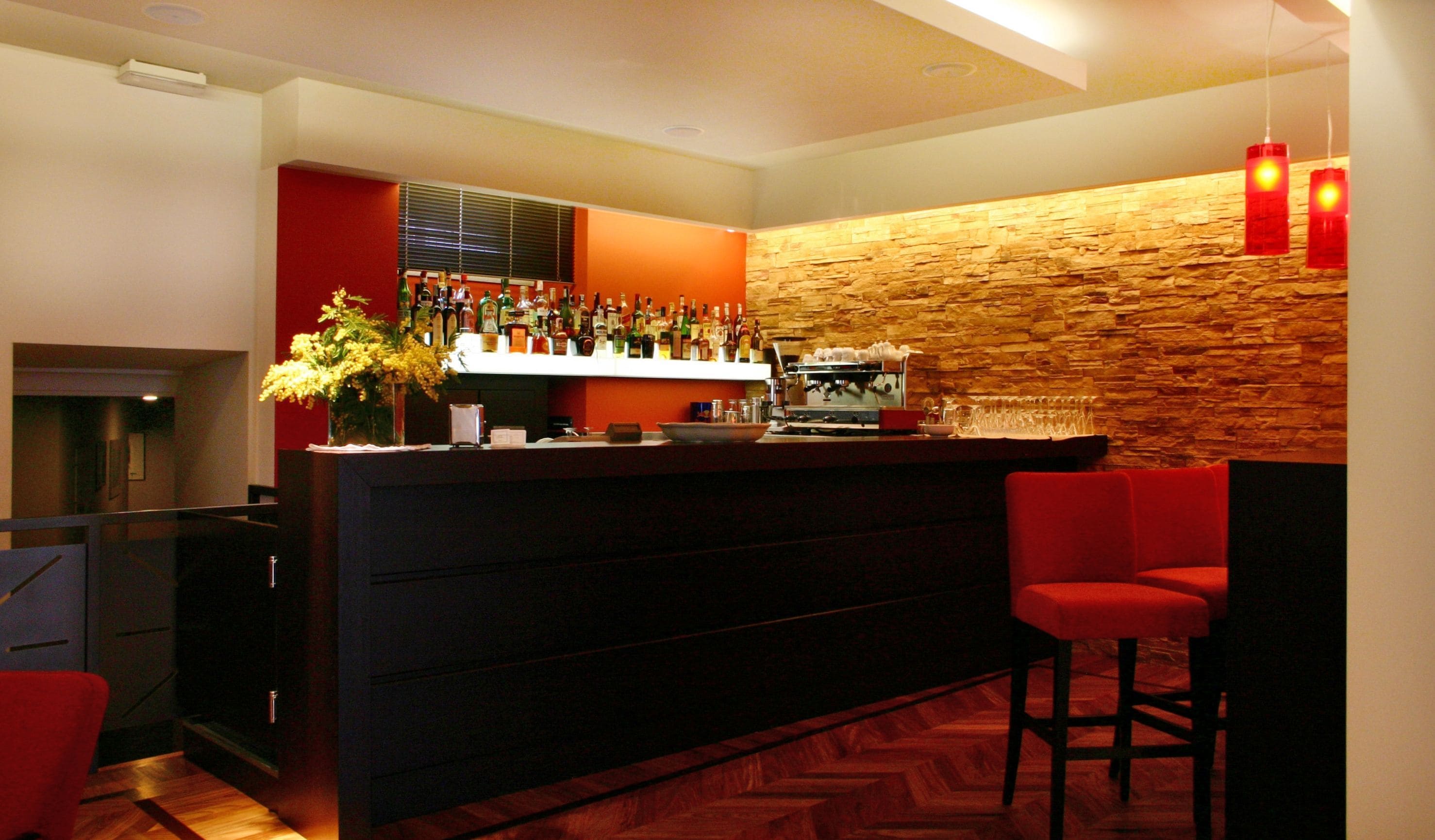 A modern bar area with a stone wall, a variety of liquor bottles, a coffee machine, and red bar stools.