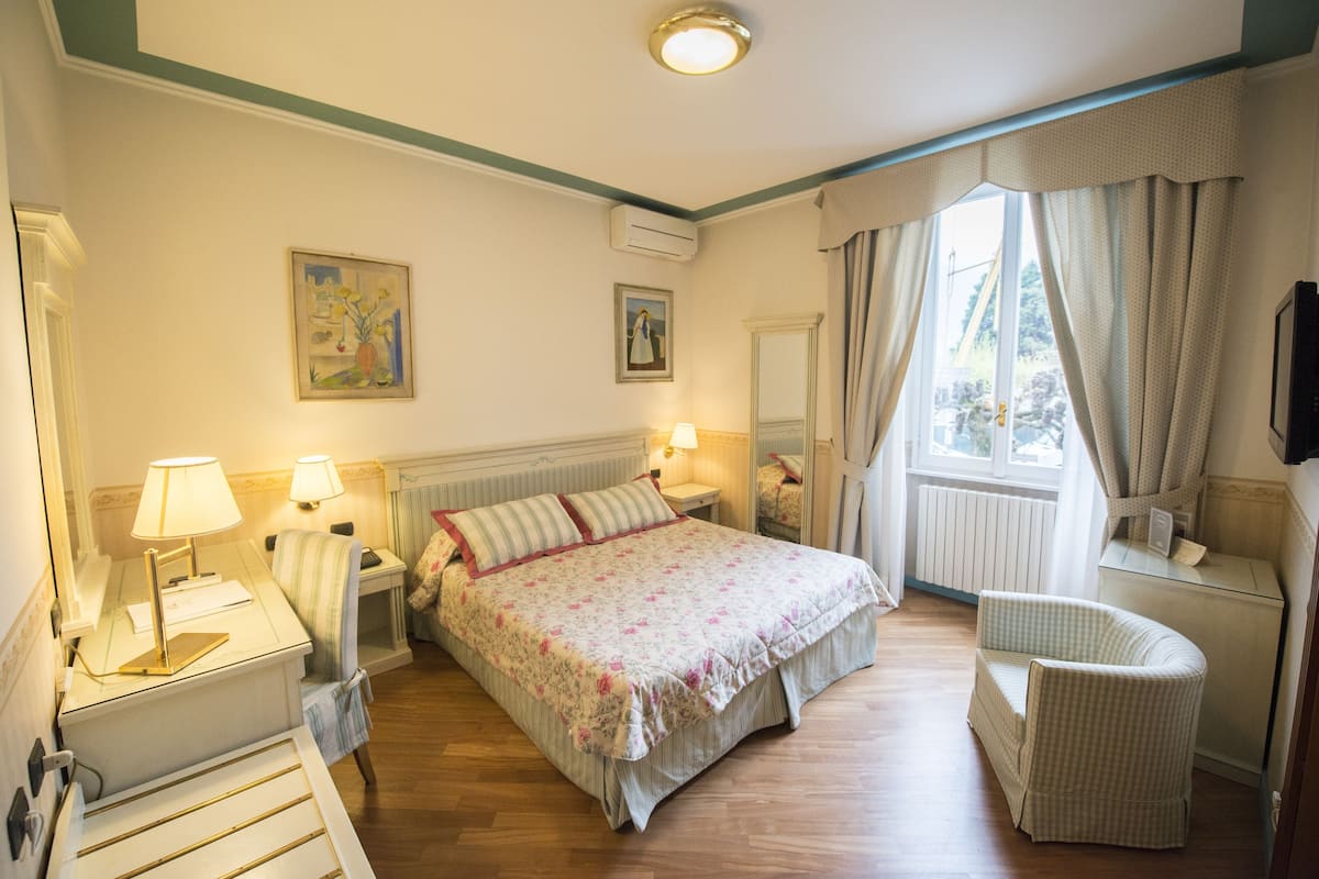 Ein Hotelzimmer mit einem Bett, zwei Nachttischlampen, einem Sessel, einem Schreibtisch mit Lampe, einem Spiegel und einem Bild an der Wand.