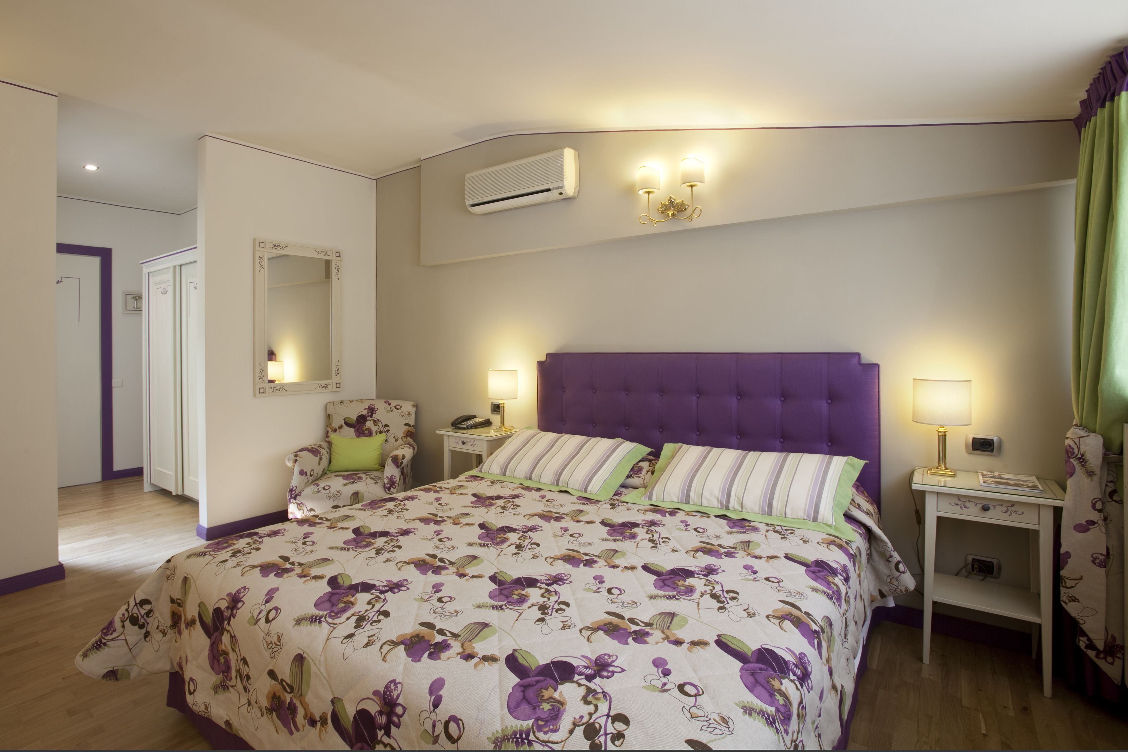 A bedroom with a large bed, a purple headboard, two bedside tables with lamps, a mirror, and an air conditioning unit.