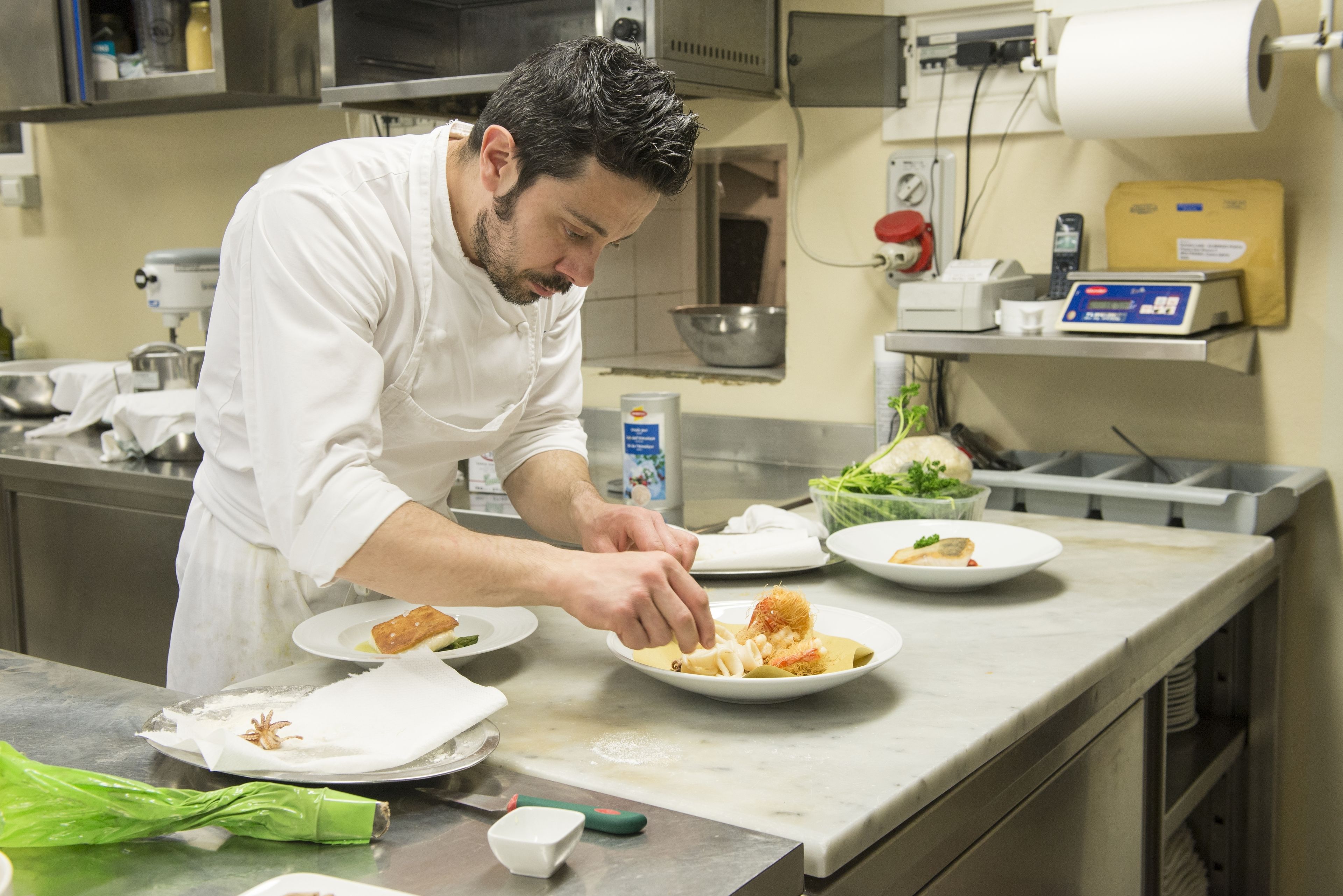 Un chef prépare des plats dans une cuisine professionnelle dotée de surfaces en acier inoxydable, de divers ustensiles de cuisine et d’une balance numérique.