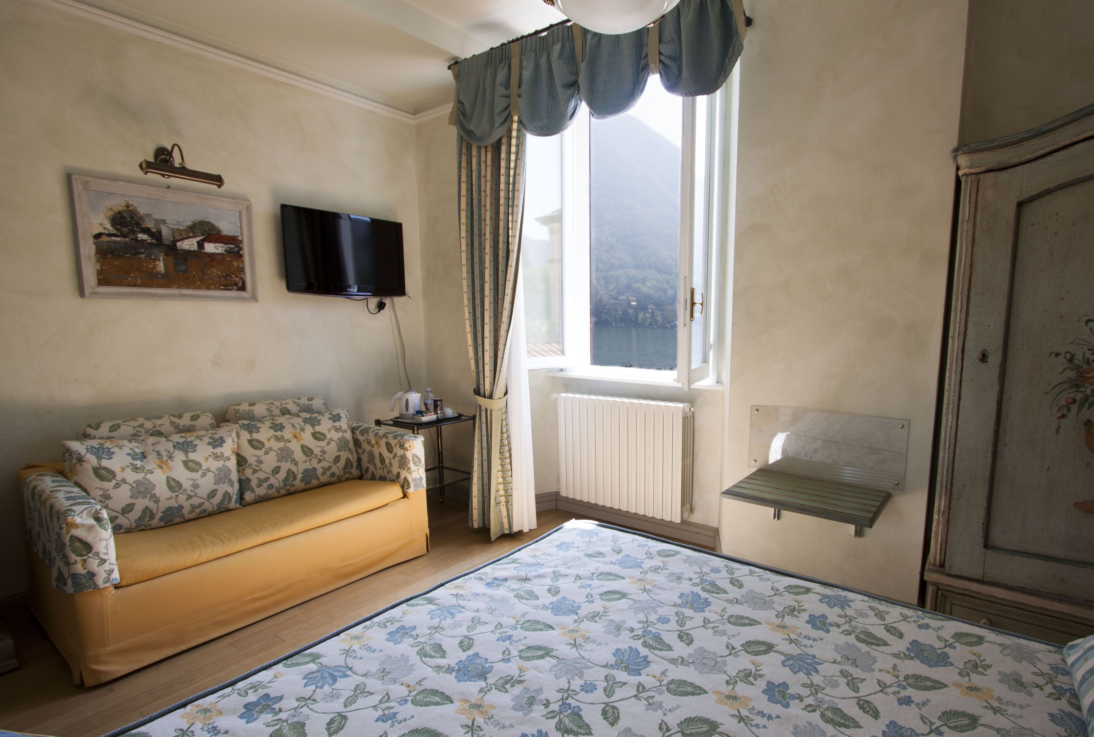 A room with a sofa, a bed with a floral bedspread, a television on the wall, a window with curtains, and a radiator.