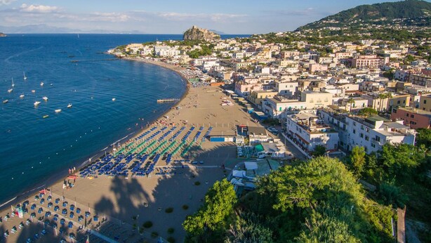 Un pueblo costero con una playa, hileras de sombrillas azules y edificios a lo largo de la orilla.