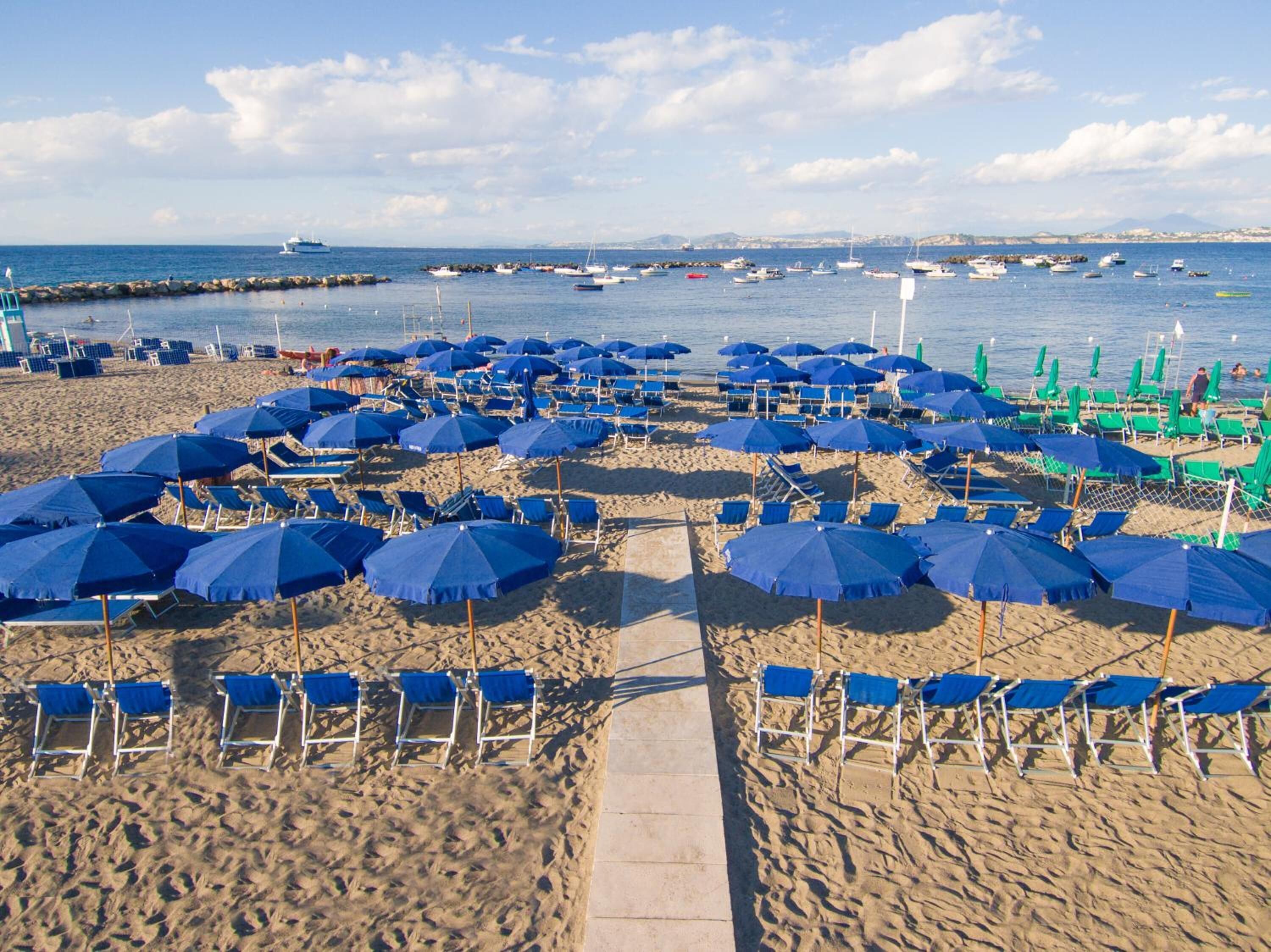 A beach with rows of blue umbrellas and chairs, a clear sky, and boats in the water.