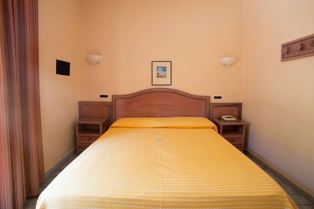 A hotel room with a wooden bed, two bedside tables, a nightstand, a framed picture on the wall, and a curtain on the left.