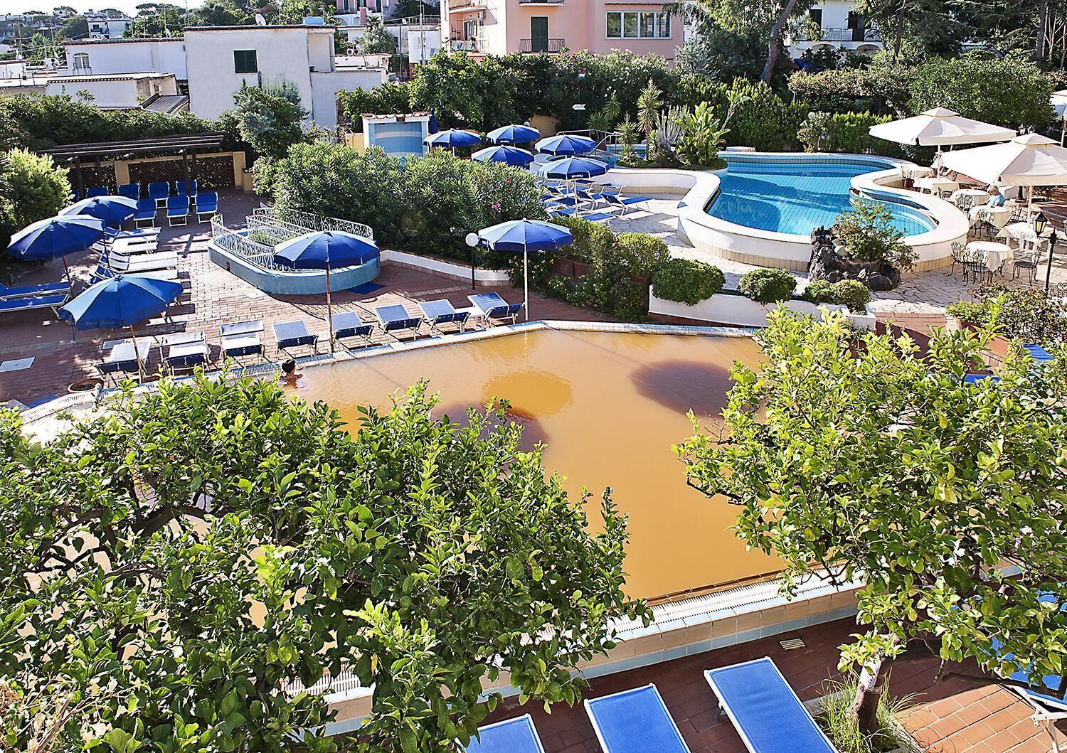 A pool area with multiple sun loungers and umbrellas, surrounded by greenery and buildings.