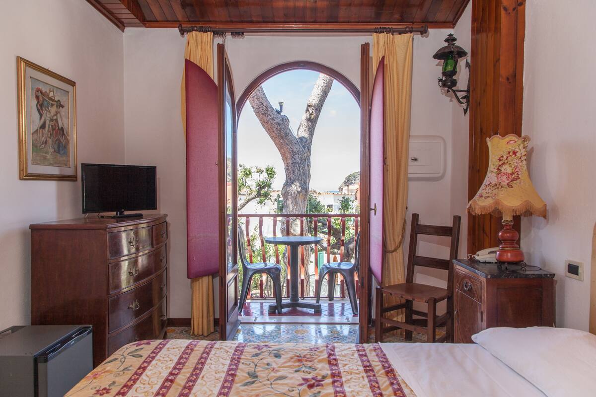 A bedroom with a bed, a dresser, a television, a table with chairs, and a balcony with a view of trees and buildings.