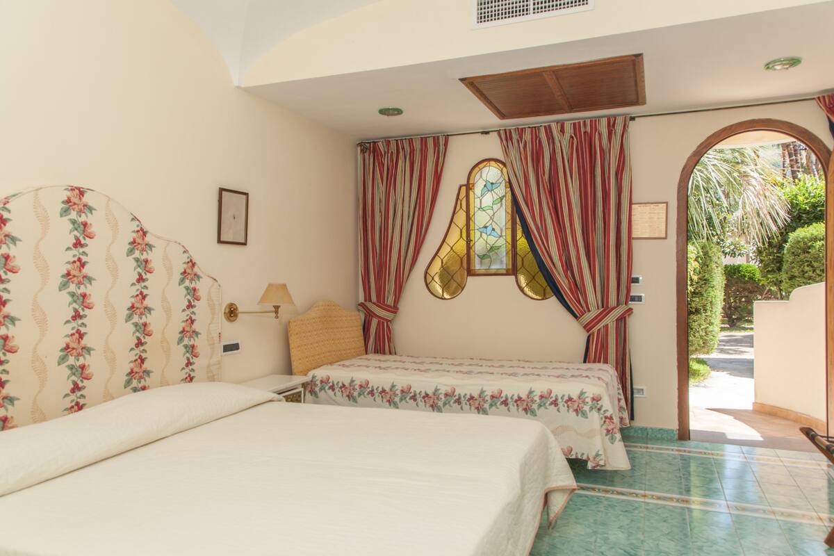 A bedroom with two beds, floral headboards, a window with curtains, and a view of greenery outside.