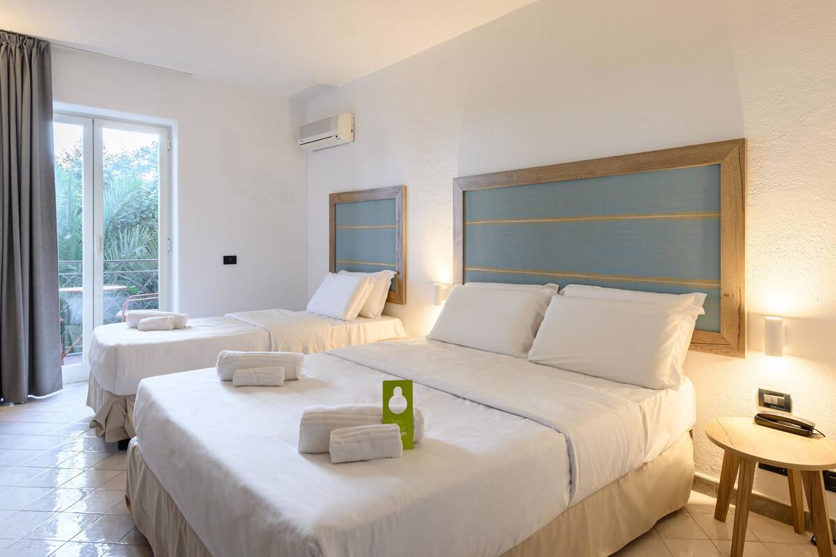 A hotel room with two beds, a small table, and a view of greenery through the window.