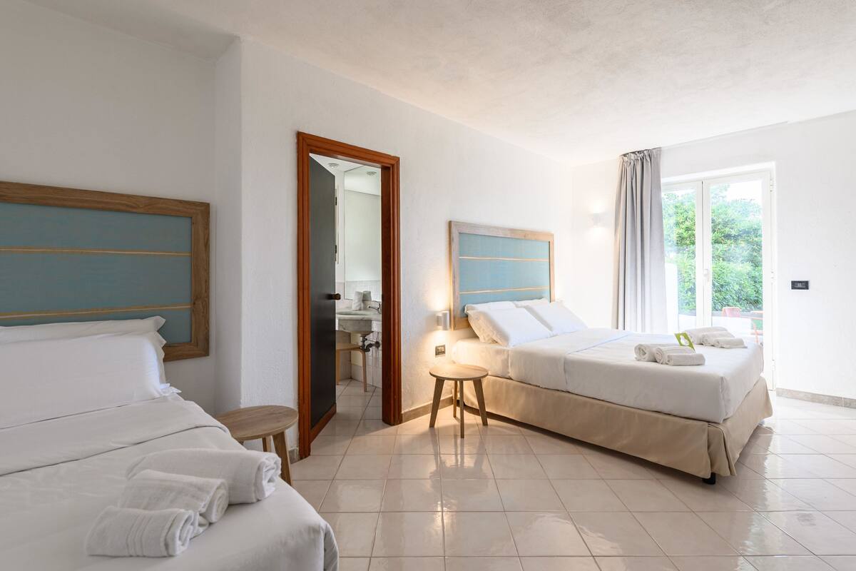 A hotel room with two beds, a bathroom, and a view of greenery.