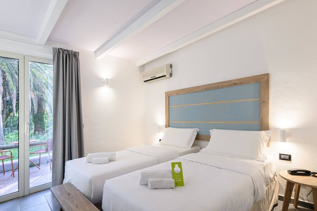 A hotel room with two beds, a balcony with a view of palm trees, and an air conditioning unit on the wall.