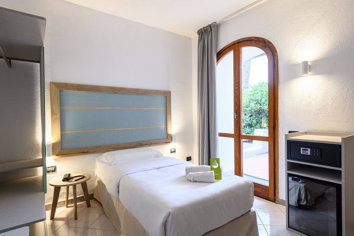 A hotel room with a bed, a small table, a desk with a computer, a large window, and a door leading to an outdoor area.