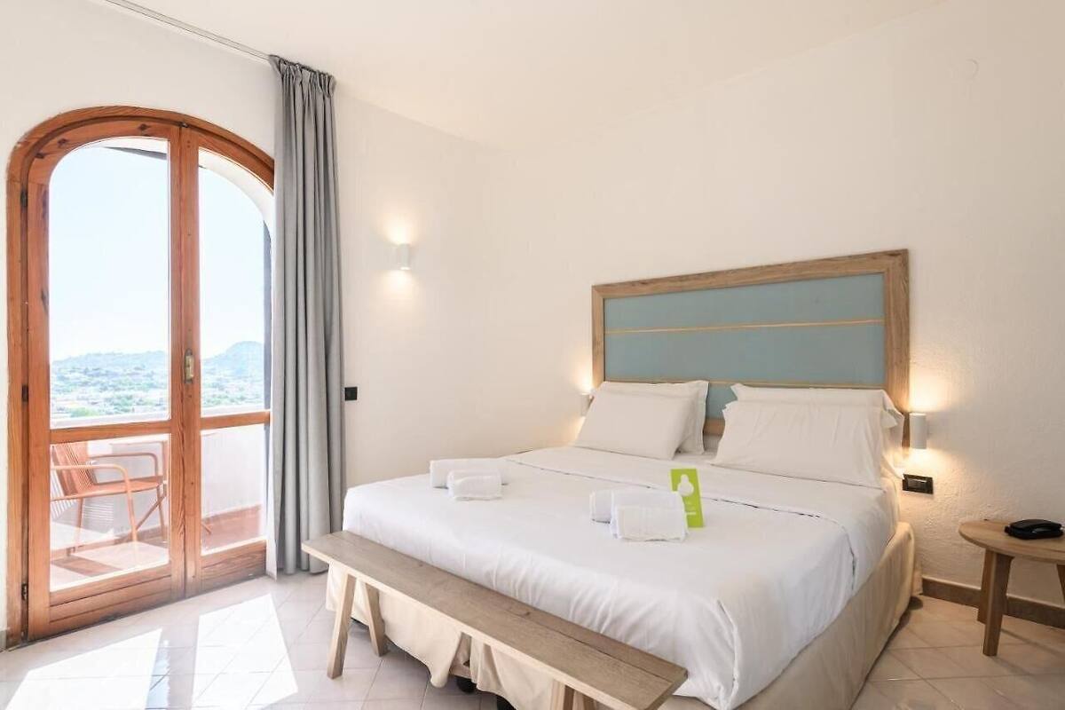 A hotel room with a large bed, a bench, a small table, and a view through a glass door.
