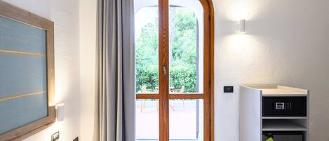 Een hotelkamer met een bed, een raam met gordijnen, een wandlamp en een klein bureau met een kluisje.