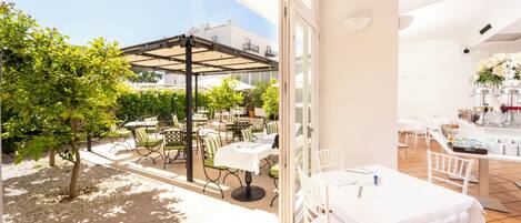 An outdoor dining area with tables and chairs, a pergola, and a view of a building in the background.