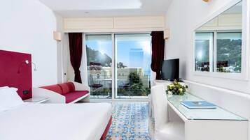 Habitación de hotel con cama, escritorio con computadora, televisión, balcón con vista a la ciudad y sofá rojo.