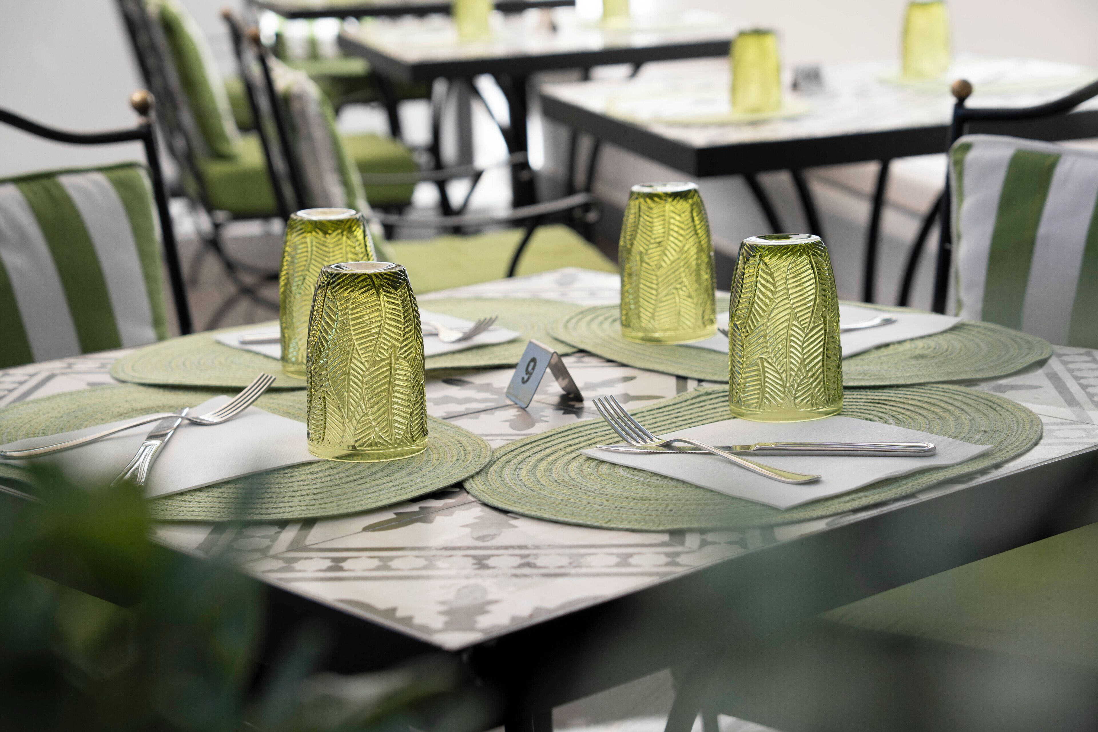 A dining area with tables set for two, each with a green placemat, a white napkin, cutlery, and a glass with a textured pattern.