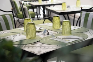 Un comedor con mesas dispuestas para dos, cada una con un posavasos verde, servilleta blanca, cubiertos y una copa con textura.