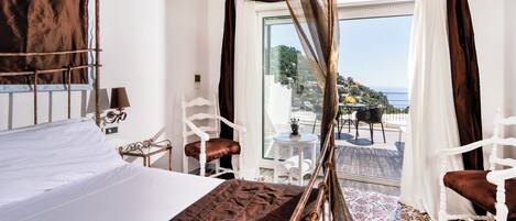 A hotel room with a bed, a chair, a table, and a view of the sea.