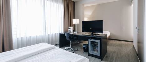 A hotel room with a bed, a desk with a computer, a TV, and a minibar.