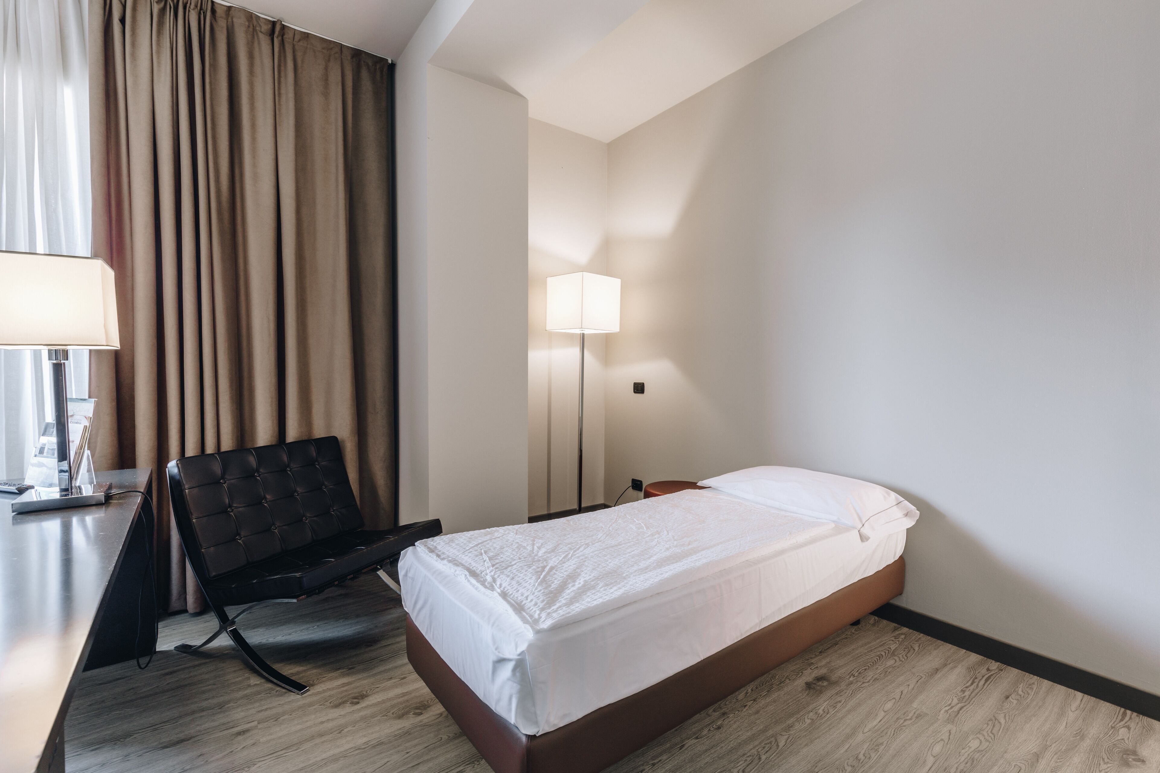 A hotel room with a bed, a desk, a chair, a lamp, and a window with curtains.