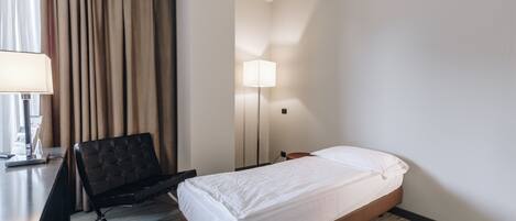 A hotel room with a bed, a desk, a chair, a lamp, and a window with curtains.
