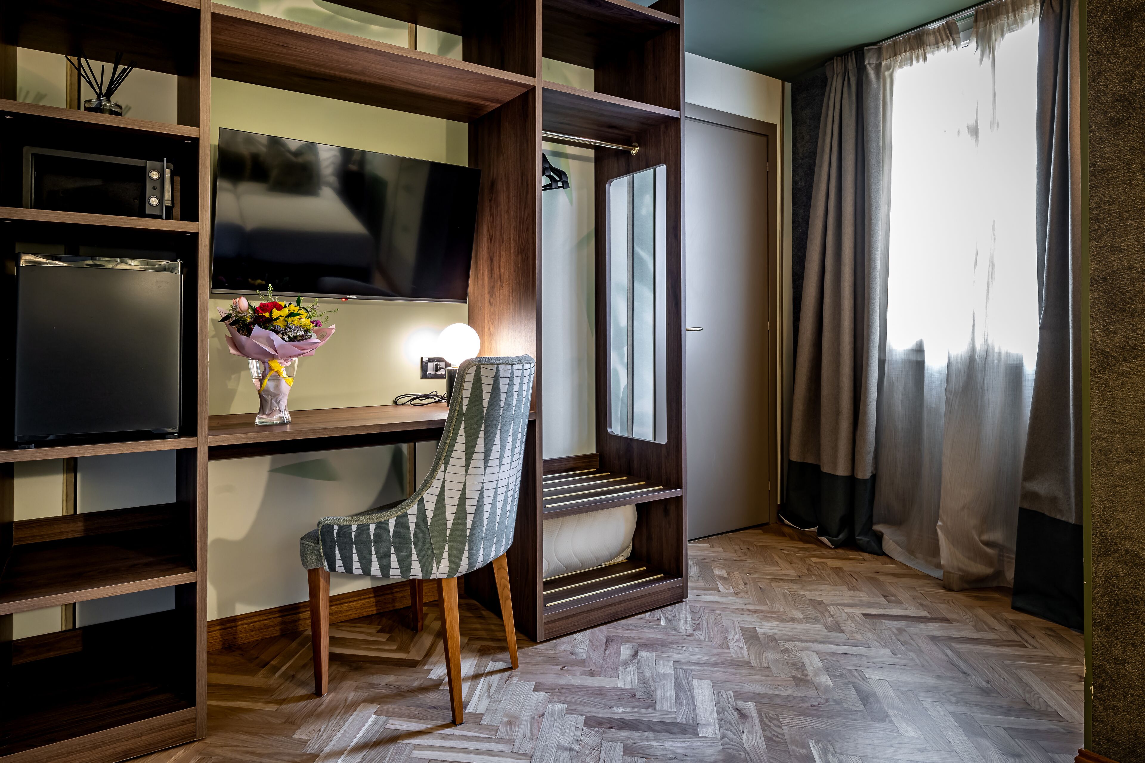 Quarto de hotel moderno com piso de madeira em padrão herringbone, uma escrivaninha com televisão de tela plana, uma cadeira, toucador com espelho e uma janela grande com cortinas.