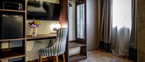 Quarto de hotel moderno com piso de madeira em padrão herringbone, uma escrivaninha com televisão de tela plana, uma cadeira, toucador com espelho e uma janela grande com cortinas.
