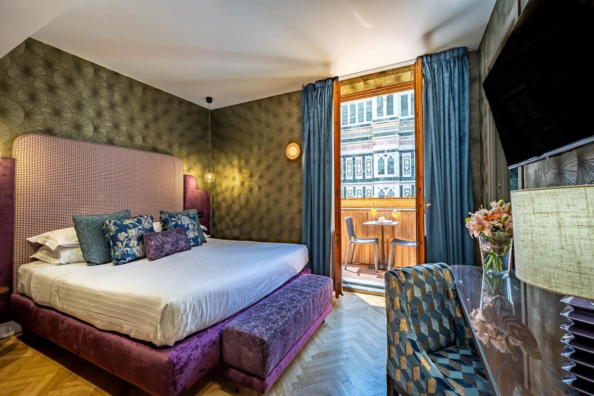 Un dormitorio con una cama grande, un banco, una silla, una mesa y vistas a un edificio a través de una ventana.
